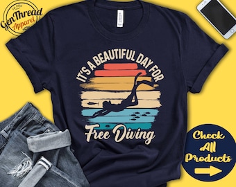 Puede incluir: Una camiseta azul marino con un gráfico de estilo vintage de una persona buceando en apnea. El gráfico presenta una puesta de sol con rayas naranjas, amarillas y rojas. El texto "It's a beautiful day for free diving" está impreso en la camiseta.