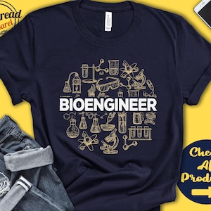 Op de afbeelding: Een marineblauw T-shirt met een wit cirkelvormig ontwerp met verschillende wetenschappelijke illustraties, waaronder bekers, reageerbuizen en een microscoop. De tekst "BIOENGINEER" is in witte letters binnen de cirkel gedrukt.