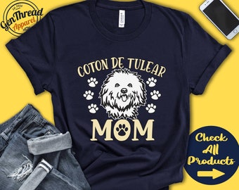 May include: A navy blue t-shirt with the text "Coton de Tulear Mom" in yellow. A white silhouette of a Coton de Tulear dog is in the centre of the shirt. There are paw prints around the dog and text.