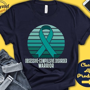 May include: A navy blue t-shirt featuring a teal green ribbon and a sunset graphic. The text "Obsessive-Compulsive Disorder Warrior" is printed on the shirt.