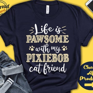 Puede incluir: Una camiseta azul marino con el texto "Life is PAWSOME with my PIXIEBOB cat friend" en blanco con un contorno amarillo. El texto está en una fuente de escritura a mano.