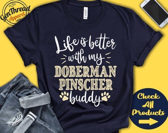 Op de afbeelding: Een marineblauw T-shirt met de tekst "Life is better with my Doberman Pinscher buddy" in wit met een gele omtrek. De tekst wordt omringd door poot afdrukken.