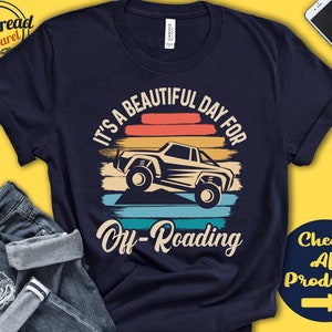 Könnte beinhalten: Ein marineblaues T-Shirt mit einer Grafik im Vintage-Stil, die ein Jeep zeigt, der durch eine Sonnenuntergangslandschaft fährt. Der Text "It's a beautiful day for off-roading" ist auf das Shirt gedruckt.