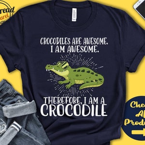 May include: A navy blue t-shirt with a white graphic of a crocodile wearing sunglasses. The text "Crocodiles are awesome. I am awesome. Therefore, I am a crocodile" is printed on the shirt.