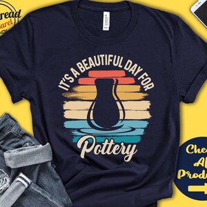 May include: A navy blue t-shirt with a retro-style design featuring a clay pot and the text "It's a beautiful day for Pottery".