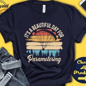 May include: A navy blue t-shirt featuring a vintage-style graphic of a paraglider flying over a sunset with the text "It's a beautiful day for paramotoring".