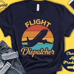 May include: A navy blue t-shirt with a vintage-style graphic of a plane flying over a sunset. The text "Flight Dispatcher" is printed in yellow below the graphic.