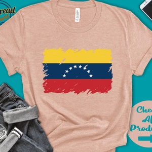 May include: A light pink t-shirt with a distressed, faded image of the Venezuelan flag. The flag is yellow, blue, and red with a white star in the center of the blue stripe. The t-shirt is a casual style with short sleeves and a crew neck.