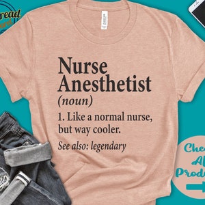 以下が含まれることがあります： 黒色の文字で「Nurse Anesthetist (noun) 1. Like a normal nurse, but way cooler. See also: legendary」と印刷された薄いピンク色のTシャツです。