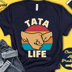 May include: A navy blue t-shirt with a vintage-style graphic of two fists bumping. The graphic is surrounded by a red, orange, and yellow sunburst. The text "TATA LIFE" is printed below the graphic.