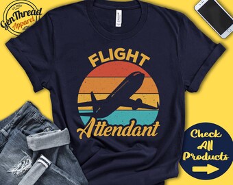 May include: A navy blue t-shirt featuring a vintage-style graphic of an aeroplane flying over a sunset. The text "Flight Attendant" is printed below the aeroplane.