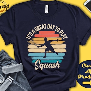 Op de afbeelding: Een marineblauw t-shirt met een vintage-stijl grafische afbeelding van een persoon die squash speelt. De afbeelding toont een zonsondergang met de tekst "It's a great day to play Squash".