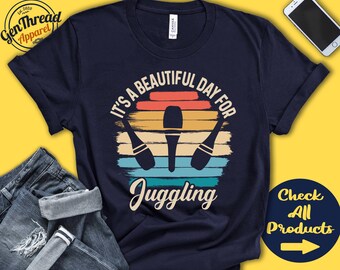 May include: Navy blue t-shirt with a retro design. The shirt features the text "IT'S A BEAUTIFUL DAY FOR JUGGLING" above a graphic of three juggling pins set against a sunset-colored background. A "Check All Products" button is in the lower right.