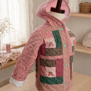 May include: A quilted hooded jacket featuring a patchwork design with pink floral fabric, dark green patterned panels, and small embroidered rabbit motifs. The jacket has a light pink interior lining, long sleeves, and four round mint green buttons.