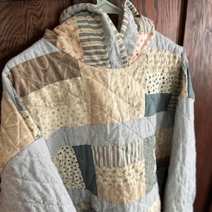 Op de afbeelding: Een patchwork hoodie in verschillende neutrale kleuren, waaronder beige, grijs en gedempte groentinten. De hoodie heeft een gewatteerd ontwerp met verschillende stofpatronen en texturen. De mouwen en de capuchon zijn effen grijs.