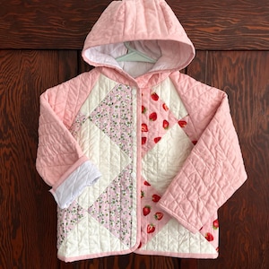 May include: A quilted hooded jacket featuring a patchwork design with solid pink sleeves and hood. The front panels alternate between white fabric, a pink floral print, and a white fabric with a red strawberry pattern, secured with white snap buttons.