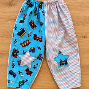 Puede incluir: Pantalones de estilo pull-on con diseño dividido, una pierna de tela azul claro estampada con trenes coloridos y un parche de estrella, y la otra pierna de tela a rayas rojas, blancas y azules con un parche de estrella a juego.