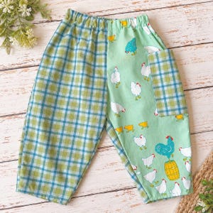 May include: A pair of pants featuring a split design with one leg in a green and blue plaid pattern and the other leg in a light green fabric printed with white chickens, yellow chicks, and a blue rooster standing on a yellow wooden barrel.