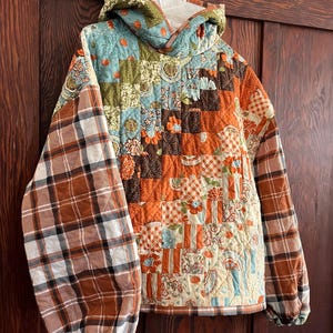 May include: A quilted jacket with a patchwork design in shades of orange, brown, blue, and cream. The sleeves are plaid, and the jacket has a hood. The jacket is hanging on a white hanger against a wooden door.