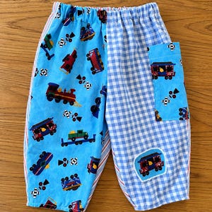 May include: Pull-on pants featuring a split design with one leg in light blue fabric printed with colorful trains and the other leg in a blue and white gingham check pattern. The pants include a matching gingham pocket and a train patch.