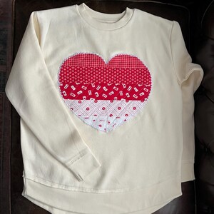 以下が含まれることがあります： クリーム色のスウェットシャツ。大きなハートのアップリケが特徴です。ハートは、千鳥格子、花柄、幾何学模様など、さまざまな赤と白の模様の生地で作られています。スウェットシャツはクルーネックで、袖口と裾はリブ編みです。