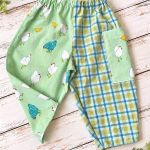 May include: These pants feature an elastic waistband and a split-leg design. One leg is light green with a repeating pattern of white and blue chickens and yellow chicks. The other leg is a blue and green plaid pattern with a matching patch pocket.