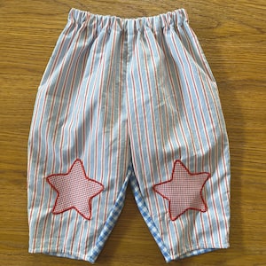 May include: A pair of pants featuring a vertical red and blue striped pattern on a white background. Each leg is adorned with a red and white gingham star appliqué. The garment has an elasticized waistband and a blue and white gingham lining.