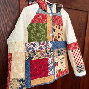 May include: A quilted hooded jacket featuring a patchwork design of various fabrics in red, blue, green, and cream. The garment includes long sleeves, a front button closure with white buttons, and a hood, showcasing patterns of paisley, hearts, and roosters.