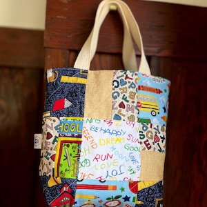 Könnte beinhalten: Eine Patchwork-Tragetasche mit cremefarbenen Griffen. Die Tasche hat verschiedene Stoffmuster, darunter Denim, Jute und Schulmotive mit Wörtern wie "Schule", "Laufen" und "Liebe".