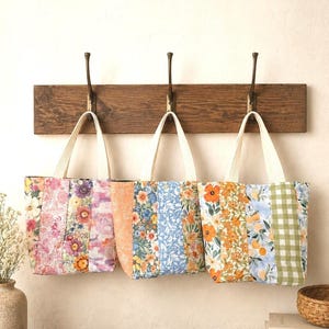 Puede incluir: Tres bolsos tote colgados en un perchero de madera, cada uno confeccionado con tiras de tela verticales con diversos estampados florales, botánicos y de cuadros en tonos rosa, naranja, azul, amarillo y verde con asas color crema.