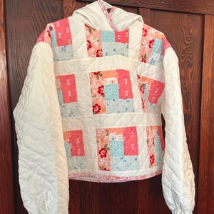 May include: A quilted hoodie featuring a patchwork design of white, light blue, and coral pink fabric panels with floral and geometric patterns. The garment has long, solid white quilted sleeves with ribbed cuffs and a matching white quilted hood.