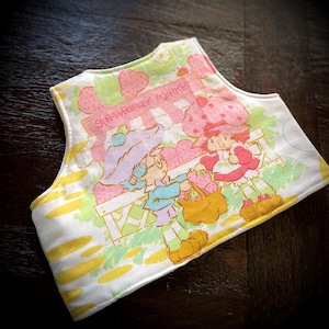 May include: A white bib with a Strawberry Shortcake design. The bib features a colourful illustration of Strawberry Shortcake and a friend at the Strawberry Market, with the text "STRAWBERRY MARKET" above them. The bib has a yellow and green striped border.