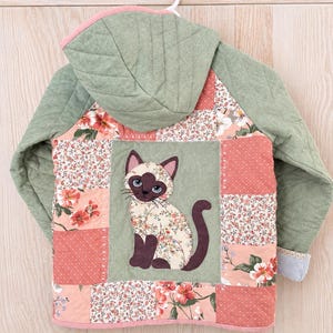 May include: A quilted hooded jacket featuring a patchwork design of floral and polka dot patterns in shades of pink, green, and white. The back panel displays a central applique of a Siamese cat with dark brown ears, face, and tail.