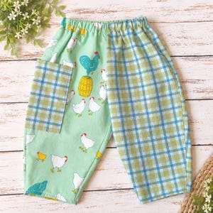 May include: Pull-on pants featuring a split design with one leg in a light green fabric printed with white chickens, a yellow barrel, and a blue rooster, and the other leg in a blue and green plaid pattern with a matching plaid patch pocket.