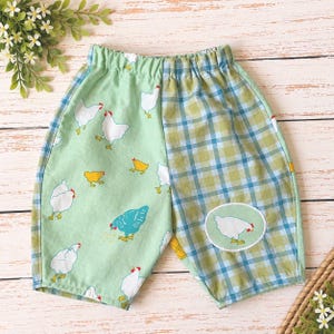 May include: Elastic waist shorts featuring a split design. The left side is light green with a pattern of white and yellow chickens. The right side features a blue and green plaid pattern with a circular patch depicting a white chicken.