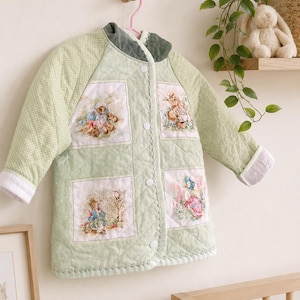 May include: A light green quilted baby jacket with illustrations of storybook characters. The jacket has a rounded collar, white buttons, and white trim on the cuffs. The jacket is hanging on a pink hanger.