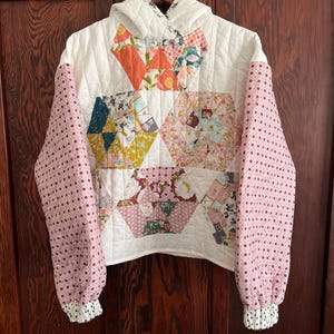 Puede incluir: Una sudadera con capucha acolchada que presenta un diseño de patchwork con varios patrones de tela florales y geométricos en tonos rosa, naranja, amarillo y blanco. La prenda tiene mangas largas con un estampado de rombos rosas.