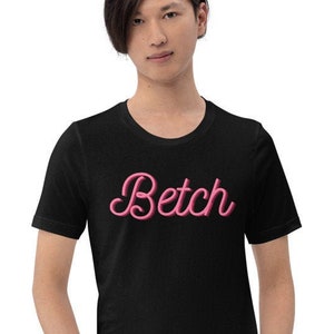 Może przedstawiać: Czarny t-shirt z napisem "Betch" w r&oacute;żowym kolorze pismem ozdobnym z przodu.