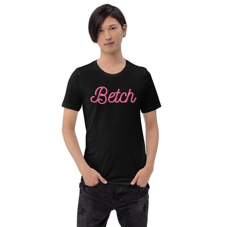 Może przedstawiać: Czarny t-shirt z napisem "Betch" w r&oacute;żowym kolorze pismem ozdobnym z przodu.