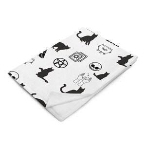 May include: White throw blanket with a black and white pattern of various witchy symbols including cats, a cauldron, a pentagram, tarot cards, a skull, and a pair of hands casting a spell.