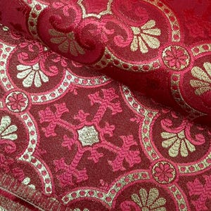 May include: A deep red fabric featuring a woven damask pattern in metallic gold thread. The design consists of repeating ornate floral motifs and cross shapes enclosed within interconnected circular and scalloped borders.