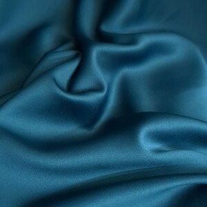 May include: A deep teal fabric with a smooth, lustrous finish and a soft, flowing texture. The material features gentle folds and curves that create subtle highlights and shadows across the surface, emphasizing the rich, saturated blue-green color.