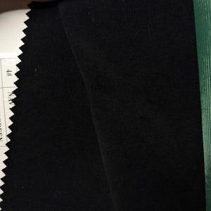 May include: A swatch of solid black fabric with a smooth, matte texture. The left edge features a zigzag pinked finish, while a white label with the text 4# NAVY is partially visible. A strip of green fabric is visible on the right side.