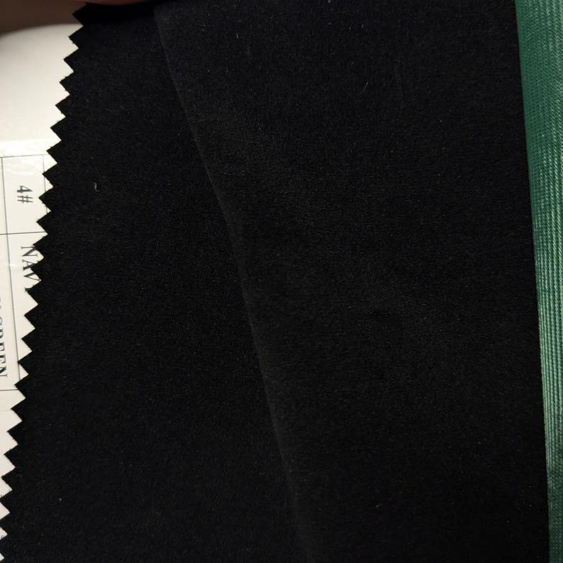 May include: A swatch of solid black fabric with a smooth, matte texture. The left edge features a zigzag pinked finish, while a white label with the text 4# NAVY is partially visible. A strip of green fabric is visible on the right side.