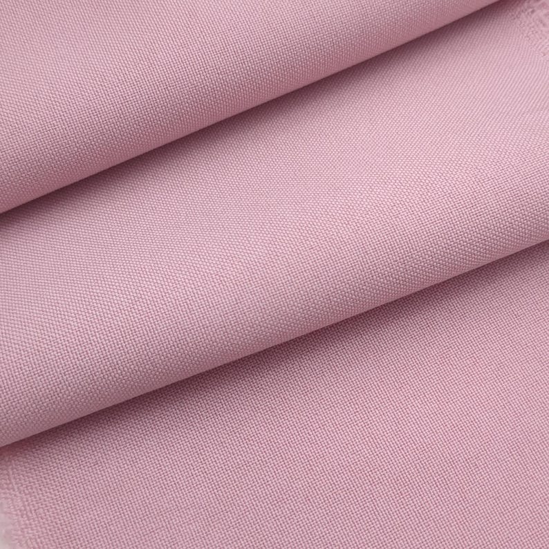 Puede incluir: Primer plano de una tela rosa claro doblada. La tela tiene un tejido fino y un acabado mate. Los pliegues crean sombras y resaltan la textura del material, de un rosa suave.