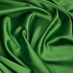 May include: A vibrant emerald green fabric with a smooth, lustrous finish. The material is draped in soft, flowing folds, creating deep shadows and bright highlights across the surface that emphasize its silky, reflective texture.