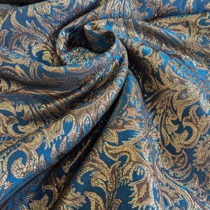 May include: A teal fabric featuring a woven metallic gold damask pattern with intricate swirling floral and leaf motifs. The material is draped to show the texture and the contrast between the deep blue-green base and the reflective gold threads.