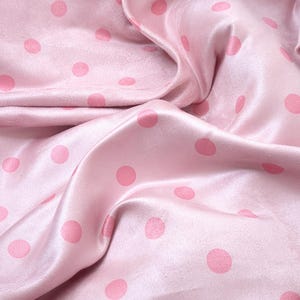 May include: A light pink fabric featuring a repeating pattern of darker pink polka dots. The material has a smooth, slightly reflective surface with soft folds and drapes that create natural shadows across the textile's surface.