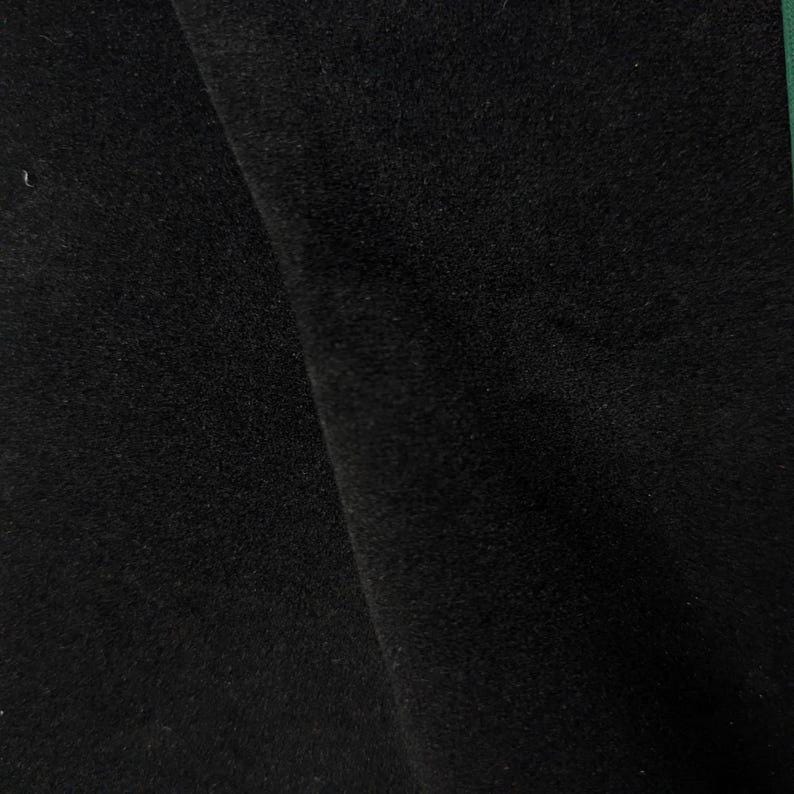 May include: A solid black fabric with a dense, soft, and slightly fuzzy texture. The material is folded, creating a diagonal shadow line that highlights the depth and consistent matte finish of the dark, uniform textile surface.
