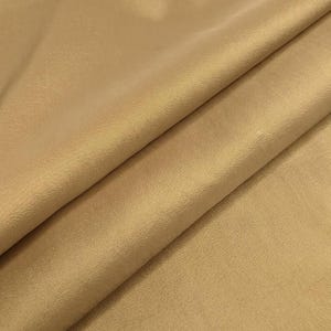 May include: A solid gold-colored fabric with a subtle, fine-grained texture. The material is draped in soft, diagonal folds, creating light and shadow across the surface. The textile has a slight sheen and a consistent, uniform metallic hue.